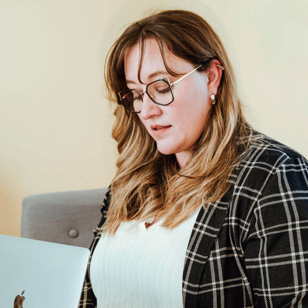 A person with long blond hair and glasses looks down while sitting indoors, holding a silver laptop. They are wearing a black and white plaid jacket over a white top. The background is neutral and softly lit.